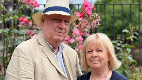 Eric and Elsie Martlew posing in a garden with a black metal fence and pink roses in the background