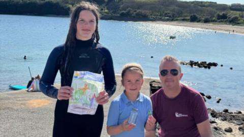 Chloe, Eliza and Matt are standing on a beach on a sunny day and there is a small bay behind them. Chloe is holding a picture drawn by Leo that was in the jam jar that Eliza is holding. Eliza is also holding the Lego spaceman. They are all smiling. 