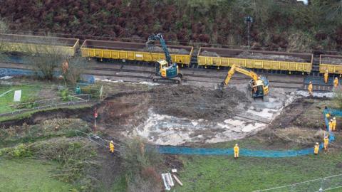 Workmen and cranes at the side of a railway track. The cranes are clearing earth from an area next to the track and loading it into yellow carriages.