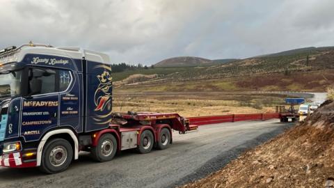 A turbine transport lorry viewed from the side with vehicles queued up behind it
