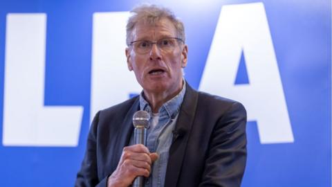 Kenny MacAskill, a man with grey hair and glasses, speaks with a microphone in his right hand, held slightly below his chin. He is wearing a dark jacket over a denim shirt in front of a blue Alba branded background.