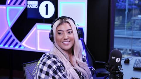 Tiffany Calver is sitting in a studio in front of a BBC Radio 1 microphone. She has headphones on is wearing a black-and-white checked jacket and has blonde hair. 