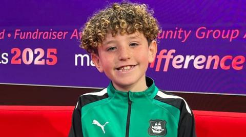 Dexter has short curly brown hair and is sitting smiling on a red sofa in the BBC Spotlight studio. He is wearing a green Plymouth FC tracksuit top. The word fundraiser and make a difference 2025 are on the screen behind him.