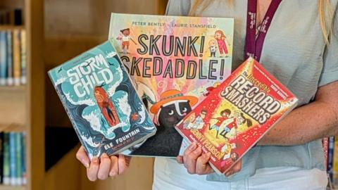A pair of hands holding three children's books with different illustrations on them.