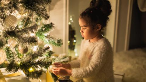 A girl with brown curly hair tied up in a bun wears a white fluffy jumper and hangs a decoration on a Christmas tree with white glowing lights on it