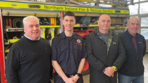 Four men of varying ages standing in front of a fire engine looking towards the camera, all wearing dark-coloured clothes. A family resemblance is visible between them.
