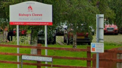 A photo of the sign outside Bishop's Cleeve Primary Academy. The sign is behind a wooden fence and there are some cars parked behind it.