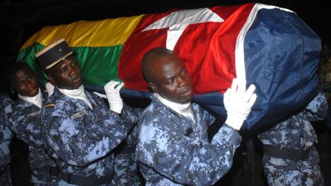 Four gendarmes, wearing blue digital camouflage fatigues and white gloves, carry a coffin bearing the Togolese flag on their shoulders