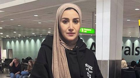 Alaa Ghalayini standing in Heathrow airport and looking straight at the camera. She is wearing a black hooded top, and a light gold coloured head scarf, gripping onto her luggage. Behind her people can be see sitting on airport seats, with a large arrival sign obscured behind a column. 