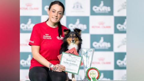 A woman in a red top wearing black leggings sits next to a brown, white and black dog and a trophy. She is holding a certificate.