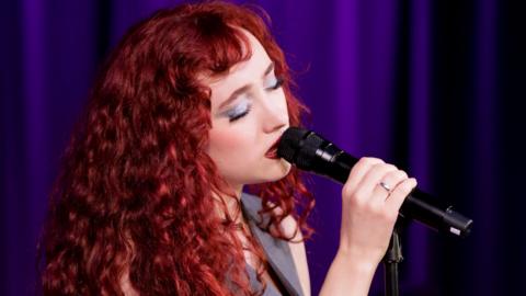 Singer Chappell Roan looking sombre as she sings into a microphone. She has red hair and pale complex. There is a ring on her wedding finger. The background is dark purple. 