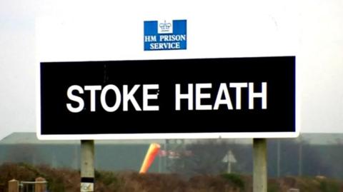 A black sign with the words Stoke Heath in white lettering. Behind them is landscape and a hedge