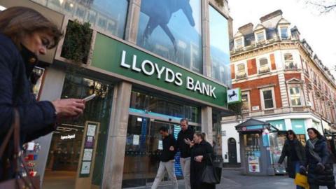 Exterior of Lloyds Bank branch with shoppers walking in front, and a woman on a smartphone closest to the camera 