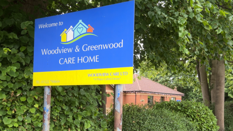 The sign outside the home. It reads "Welcome to Woodview & Greenwood CARE HOME". This is on a blue background which takes up most of the sign. There is a yellow strip at the bottom with the phone number and the company name "Woodview Care Ltd"