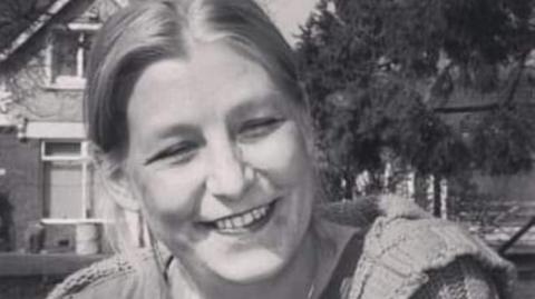 Dawn Sturgess - a black and white image, where Dawn is sat outside with a house and trees behind her. She has light hair and is smiling into camera.