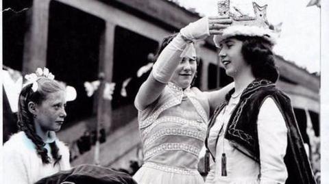 A black-and-white image of a woman being crowned by another woman. She is wearing a black cape and white dress; the woman placing the crown on her head is wearing a white dress. A young girl stands by with a cushion from which the crown has just been lifted - she is also wearing a white dress.