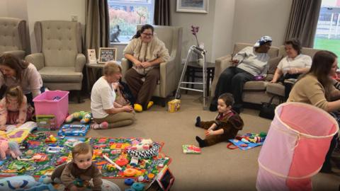 Living room area in the care home, with children and toys on the floor and parents and residents looking on from chairs
