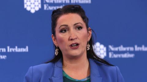 Emma Little-Pengelly. She has long dark hair partly tied back, wearing a blue blazer and pearl drop down earrings. A blue background is behind her with Northern Ireland Executive branding on it. She is standing in front of a microphone.