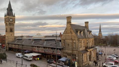 View of Darlington market hall in the town centre. It has a Gothic looking two-storey building at one end, then arched windows along a promenade leading to a large Gothic church tower on the other.