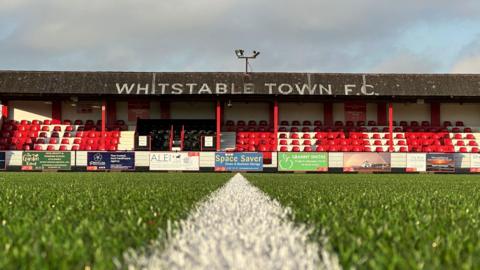 A photograph of a football stand with WHITSTABLE TOWN F.C. written on the roof. The photo is taken from grass level on the other side of the pitch