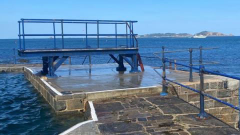The diving board is pictured at high tide against a blue sky with Herm in the background. 