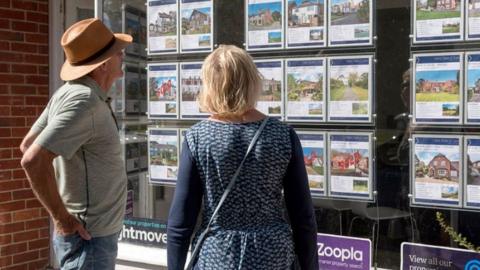 A man and a woman on a street look at houses on the market in an estate agent's shop window