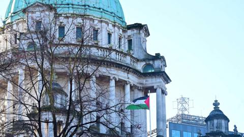 The Palestine flag is flapping in the wind outside a baroque revival style building. Several pillars decorate the building. The roof is a green dome. The sky is light blue. A tree is bare of leaves.