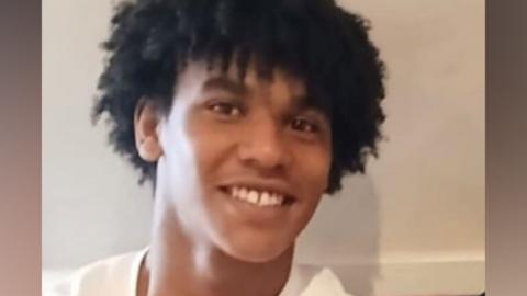 Devon Simmonds-Caines, with black mid-length curly hair and wearing white top, smiles at the camera