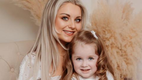 Hollie pictured on a cream chair with her mum Ann, the photo is professional. Hollie has long curly red/brown hair and is wearing a blue and white dress with a white cardigan. Ann has long blonde hair and is holding Hollie. 