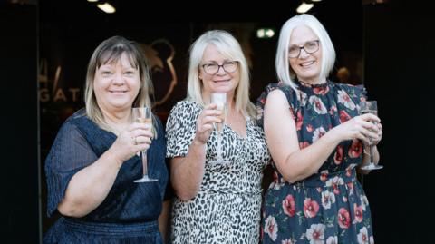 Kathy, Sarah and Debbie all holding a glass of champagne, smiling at the camera 