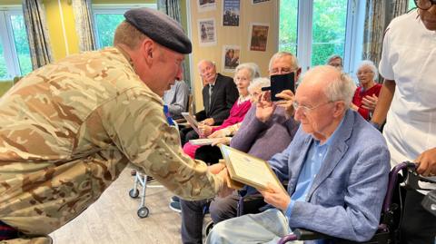 A person in military uniform presents a framed certificate to an elderly man sat in a wheelchair. Other elderly people nearby also hold certificates. The room features large windows and pictures on the walls.