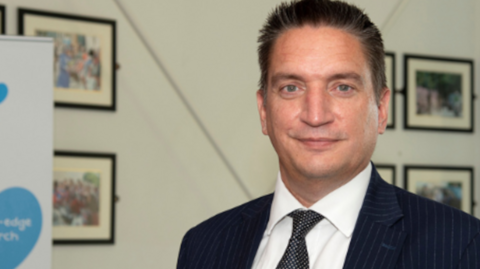 Adam Sewell-Jones a man standing in front of a wall with framed photographs on it. He is looking directly at the camera and smiling and is wearing a black pin striped suit jacket, white shirt and black patterned tie.