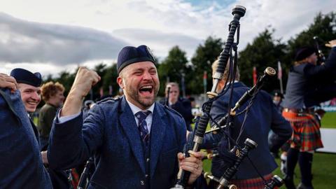 A piper from the Simon Fraser University pipe band celebrating their win. His mouth is open in a wide smile and he is punching the air. He is wearing a traditional bagpiper outfit with hat and is holding his bagpipes. There are other pipers behind him.
