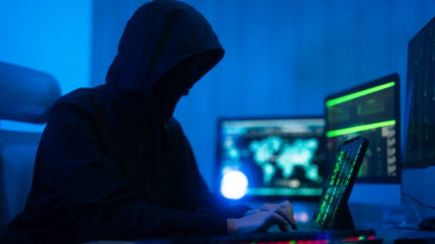 A person wearing a hooded coat is typing on a computer. They have multiple screens open and appear to be doing something covert. The room is illuminated with a dark blue light.
