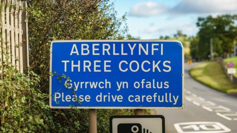 A road sign as you enter Three Cocks village warning people to drive carefully in English and Welsh.

