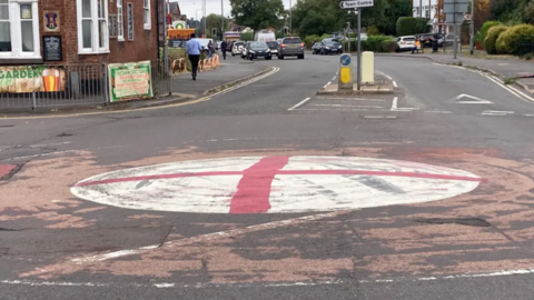 A red cross painting on a white mini roundabout, at the junction of a road that has a sign on it, with Town Centre on it. A pub can also be seen, with pedestrians and cars in the background.