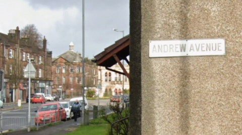 A road with cars parked on each side in the distance. In the foreground, a white sign on a grey wall reads Andrew Avenue