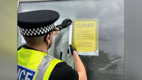 Police officer in green vest putting a cello tape on the closure notice on the door 