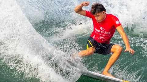 Young male surfer in a red top and black shorts making a clean cutback on a wave with copious spray.