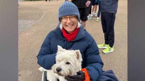 A woman wearing a navy hat, coat, red scarf and leather gloves sitting in a wheelchair smiling at the camera. She is holding a small white dog on her lap. 