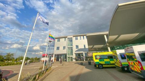 Darent Valley Hospital in Dartford, England, with people visible in the background and the NHS and Gay Pride flags outside.