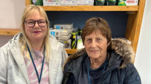 A woman with blonde shoulder-length hair and glasses, wearing a pink striped jumper and a grey coat, stands next to a woman with short brown hair who is wearing a navy jumper and navy coat. Behind them, wooden shelves hold household goods.