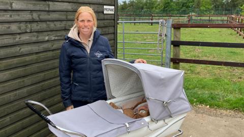 Ros Canter pictured with a pram and baby outside a stable. She is wearing a blue padded jacket and is standing next to a field enclosure.