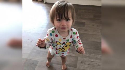 A picture of a smiling Bronson Battersby who has dark eyes and dark hair. The toddler is standing on a grey wooden floor wearing a polka dot top with a Pudsey bear design on the front.