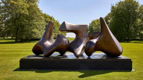 Large curved bronze sculpture by Henry Moore on a plinth, surrounded by open grass and trees in bright sunlight