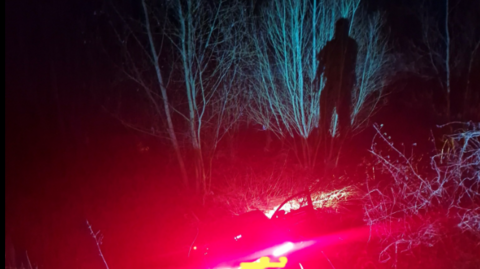 A police officer stands by the side of the river in the dark, lit up by a red light from the police car