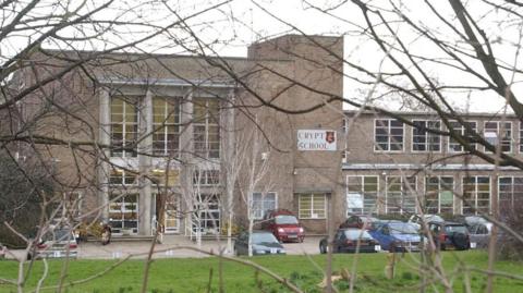 A brown-brick two-storey school, with flat roofs. There is a small car park in front, and a large sign reading "Crypt School" halfway up the building. Branches are in the foreground.