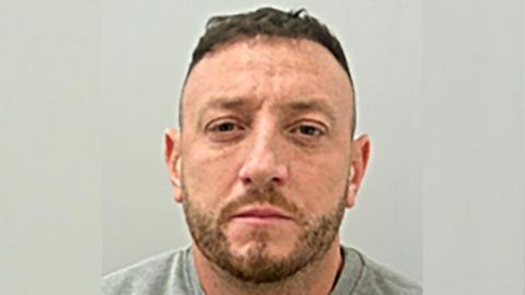 Jake Balmforth is looking straight at the camera in this police mugshot. He has short brown hair and stubble and is wearing a grey top.