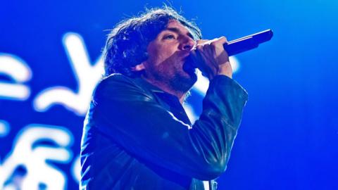 Snow Patrol singer Gary Lightbody sings into a microphone on stage wearing a blue jacket. Behind him is a blue screen with white letters.