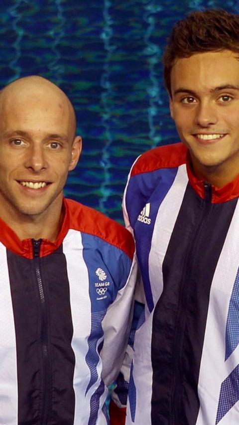 Pete Waterfield stands next to Tom Daley in front of a swimming pool. They are both wearing Team GB Olympics jackets.